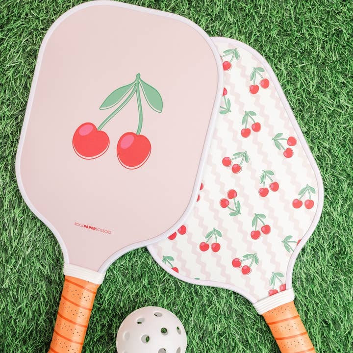 Two pickleball paddles with cherry designs on a grass background
