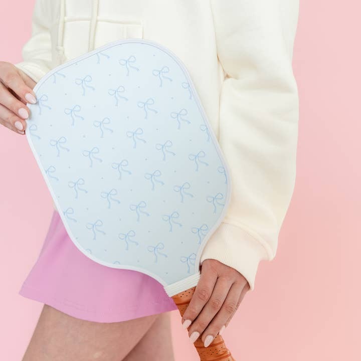 Light blue pickleball paddle with blue bow patterns held by a person against a pink background