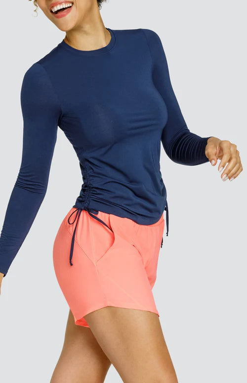 A woman wearing a navy blue long sleeve top with a crew neck and drawcord sides, paired with coral colored shorts.