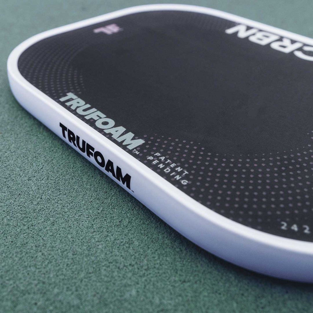 A black and green CRBN Tru Foam Genesis Hybrid 14mm paddle with a foam core, alongside a black carrying case. The paddle features geometric patterns and branding details in white and green colors.