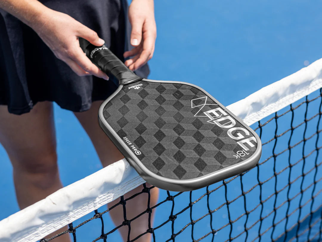 A close-up of a black and gray Diadem Edge 18K Speed Pro pickleball paddle with a geometric pattern, being held by a person with a tennis racket in the background.
