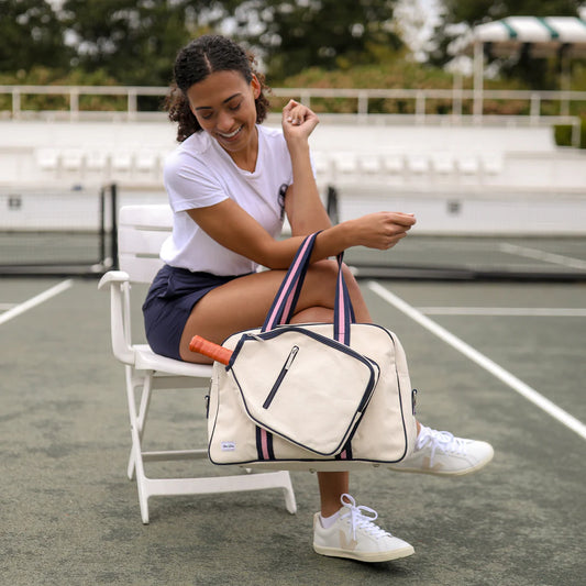 A person sitting on a chair with a pickleball tote bag on their lap. The bag is white with red and blue accents, and it has a paddle-shaped pocket visible.