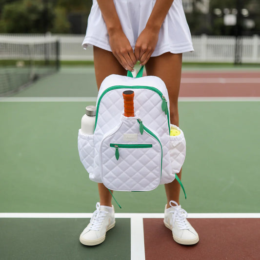 A person holding a white and green Ame & Lulu Pickleball Time Backpack with a quilted design, featuring a paddle shaped pocket, on a pickleball court.
