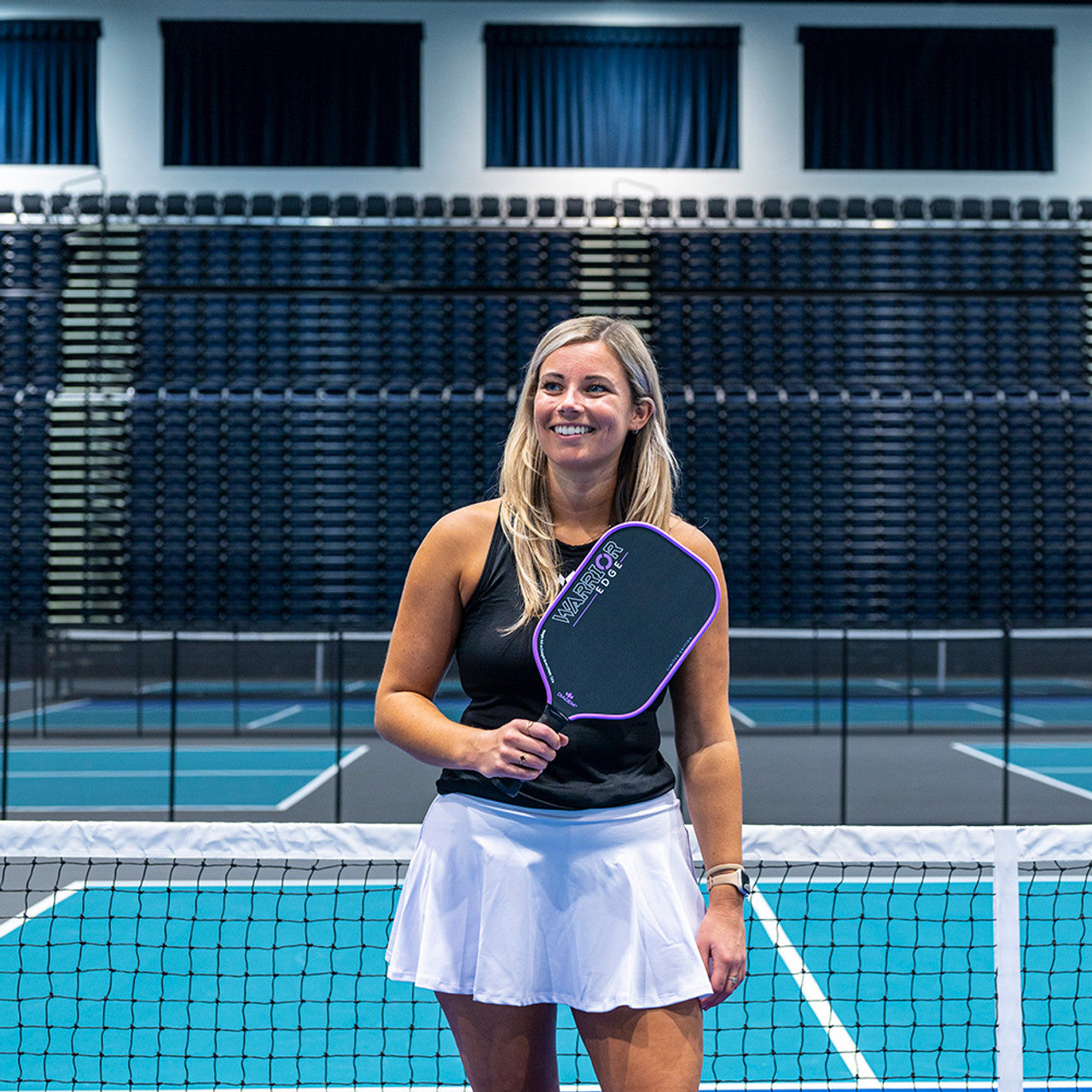 A black and pink Diadem Warrior Edge paddle with a carbon fiber pattern and the brand name etched on the surface. Held by a woman on a pickleball court