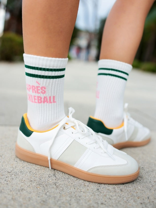 White sneakers with green accents worn with white socks featuring pink text on a blurred outdoor background